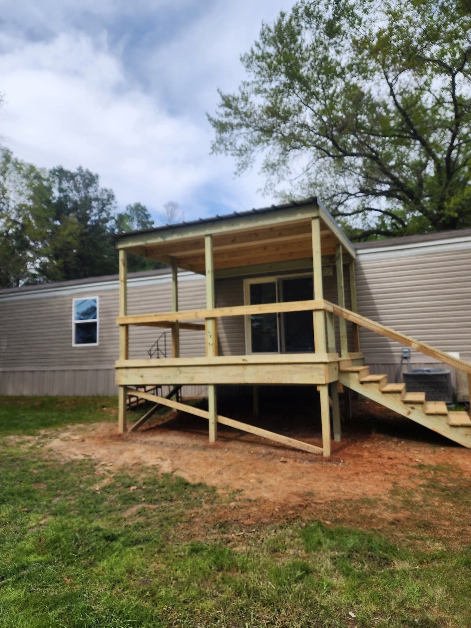 Covered wooden porch and ramp attached to a beige mobile home, with steps and grassy yard.