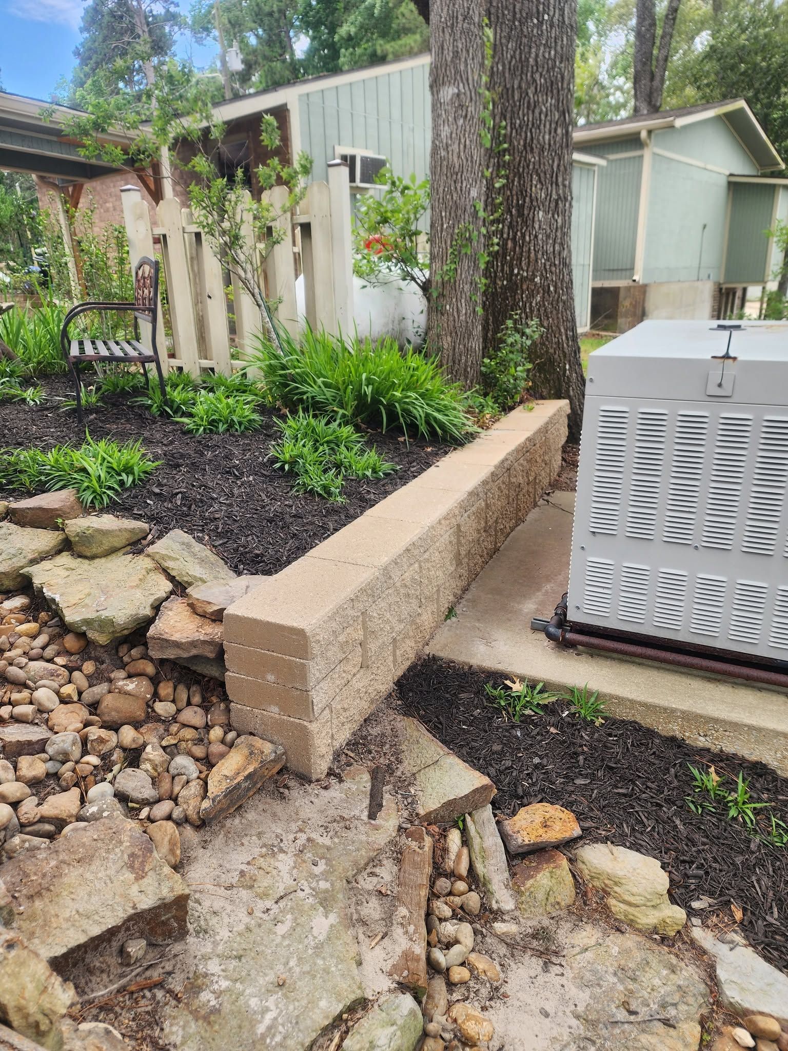 Backyard garden with stone retaining wall, trees, mulch, and a white outdoor unit beside a shed