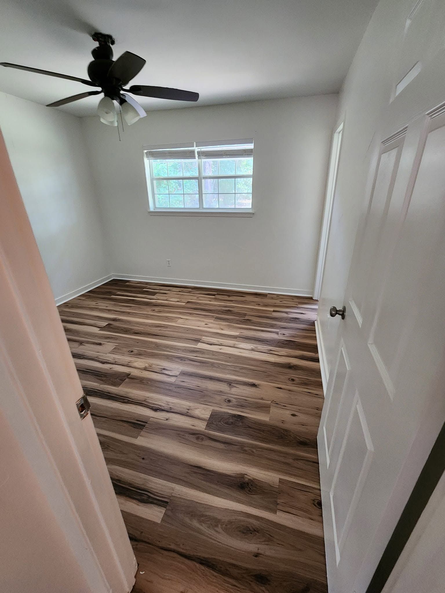 Empty small bedroom with white walls, wood-look flooring, ceiling fan, and a window
