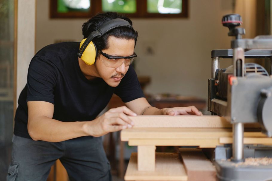 Man using earmuffs measures wood in a workshop beside a woodworking machine