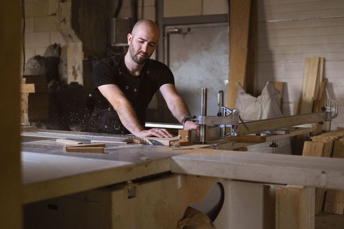Woodworker in a workshop measuring a wooden board on a workbench, with tools and sawdust nearby