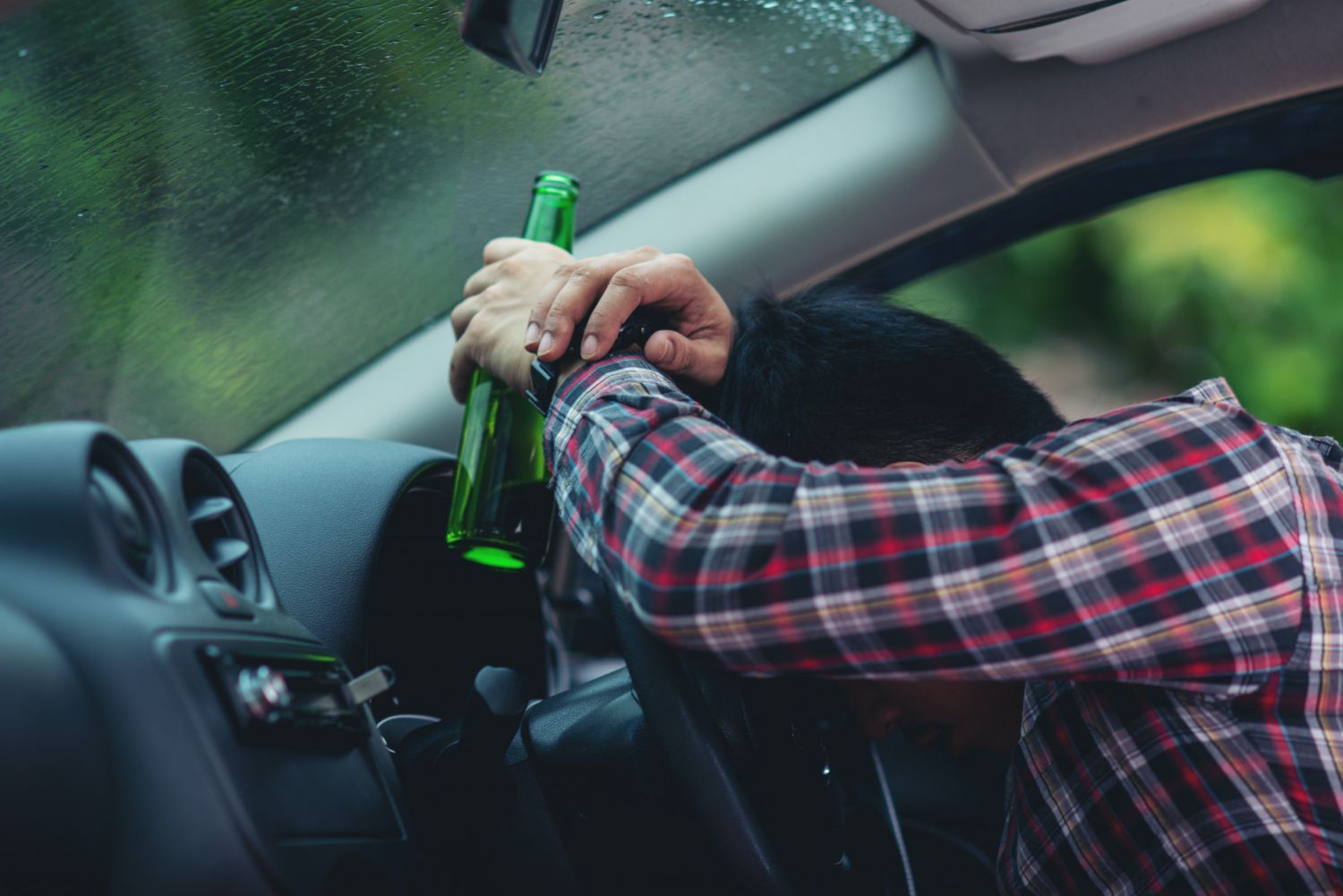 A person wearing a plaid shirt rests their head on a steering wheel while holding a green glass bottle inside a car.