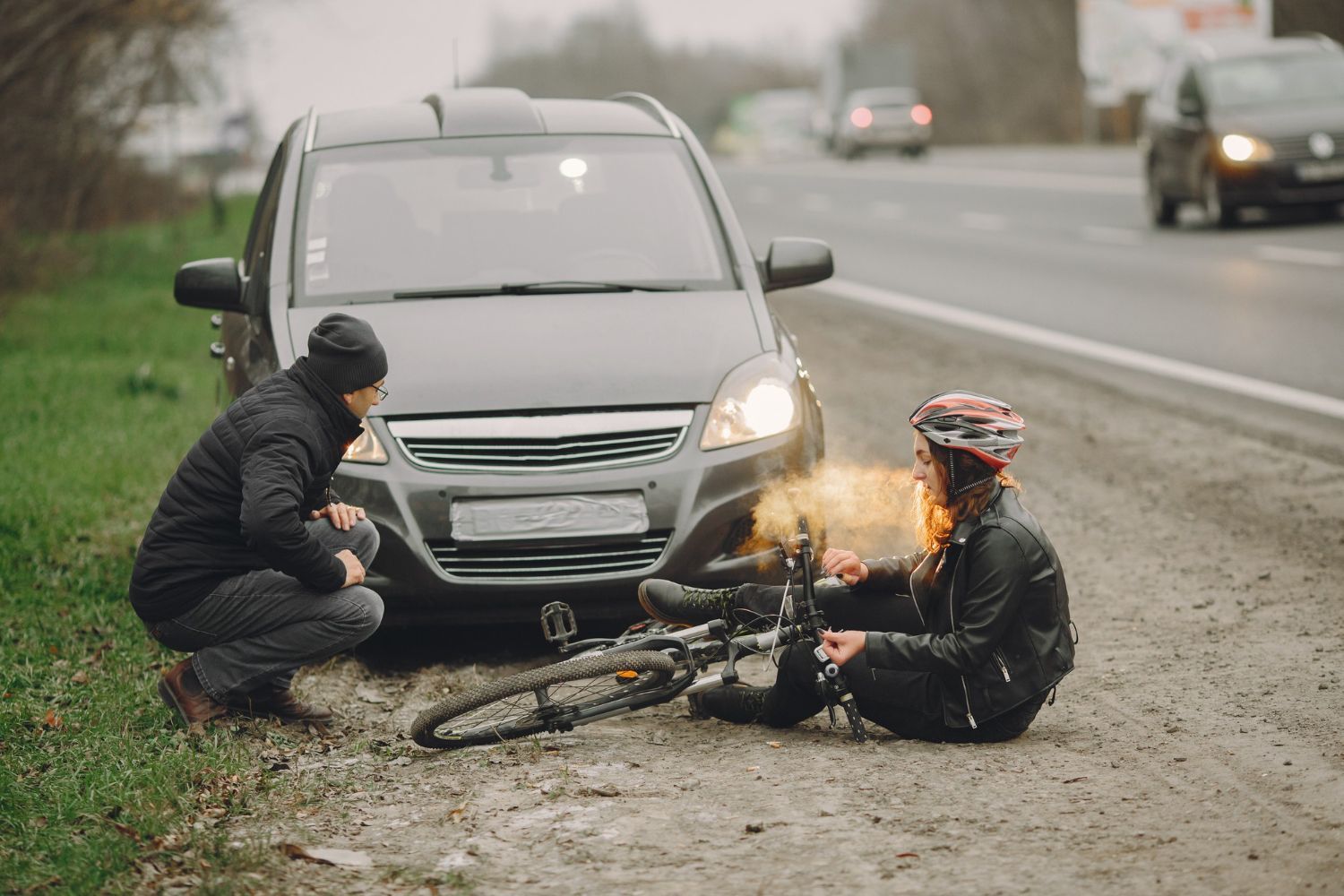 A person crouching to help a cyclist sitting on the gravel shoulder in front of a car parked on the roadside.
