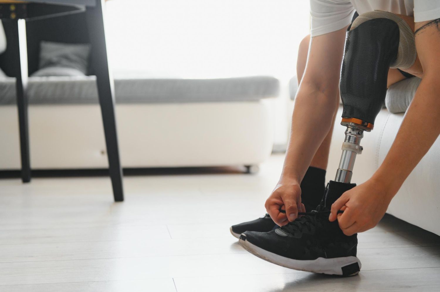 A person wearing a prosthetic leg sits on a couch and ties the laces of a black athletic shoe.
