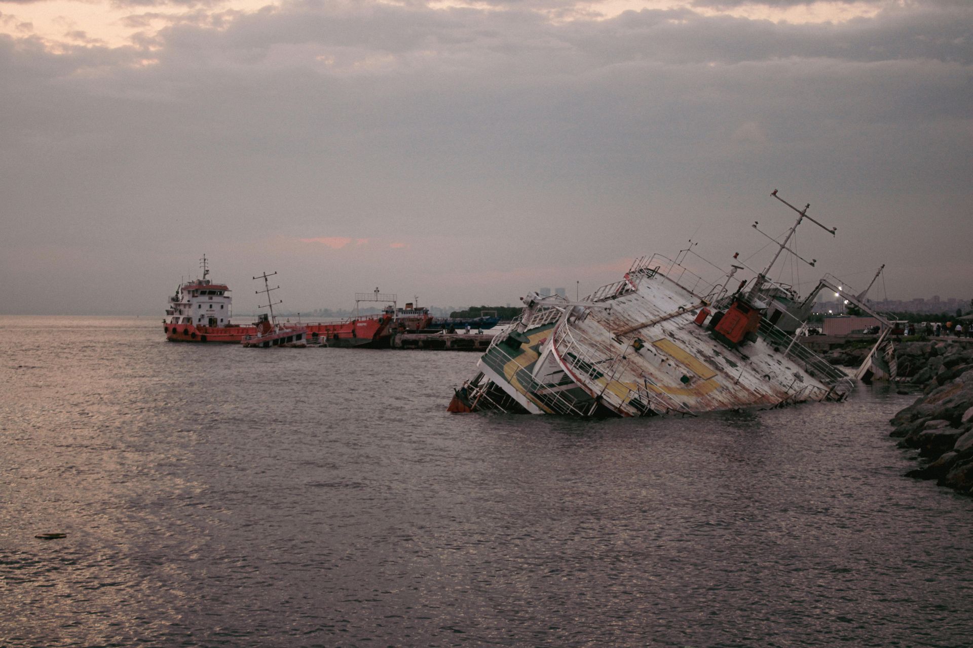A partially submerged ship tilted on its side near a rocky shoreline, with another vessel visible in the background.