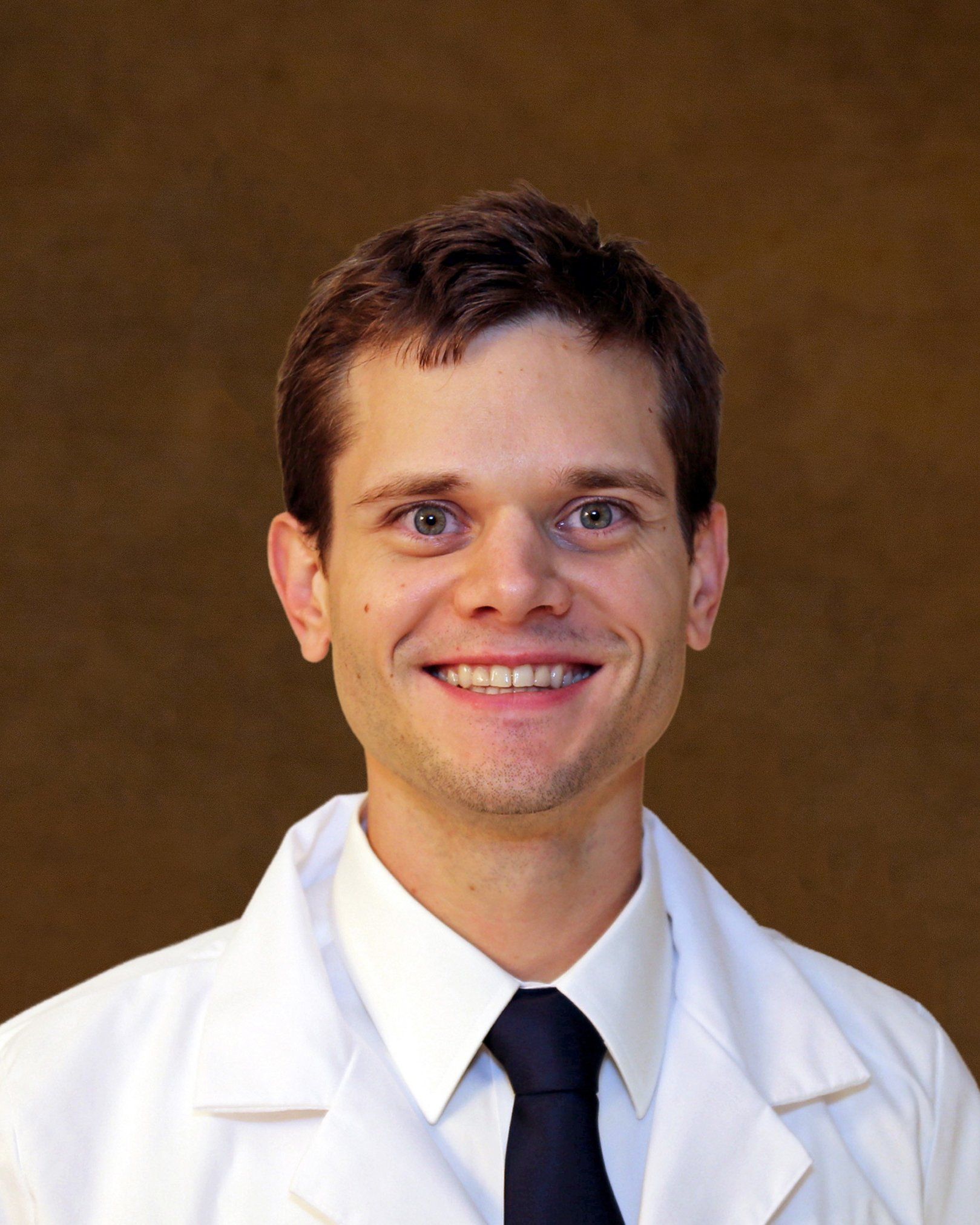Man in white lab coat smiles at the camera; dark tie and neutral background.