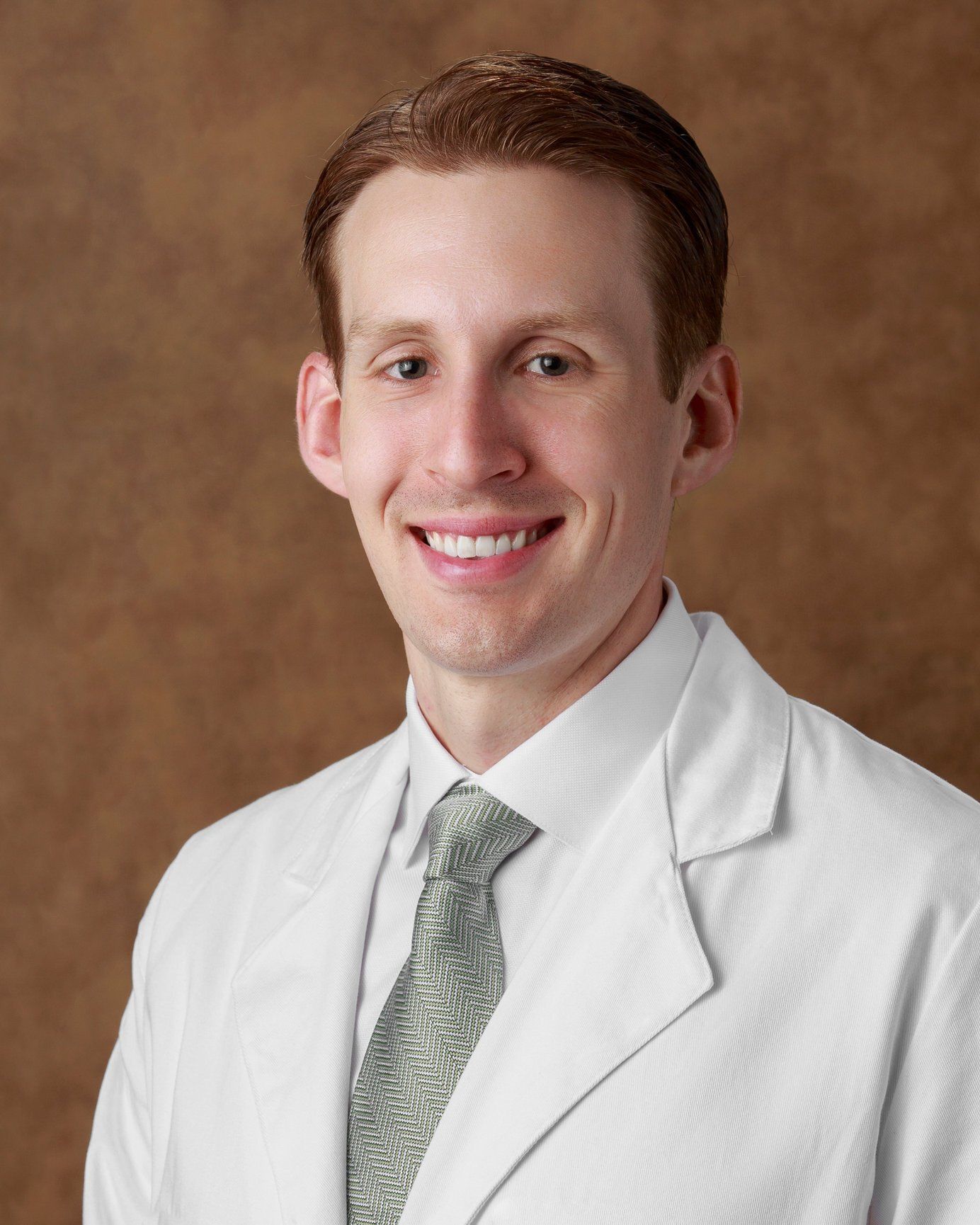 Smiling man in white lab coat, light-colored tie, brown hair, against a brown backdrop.