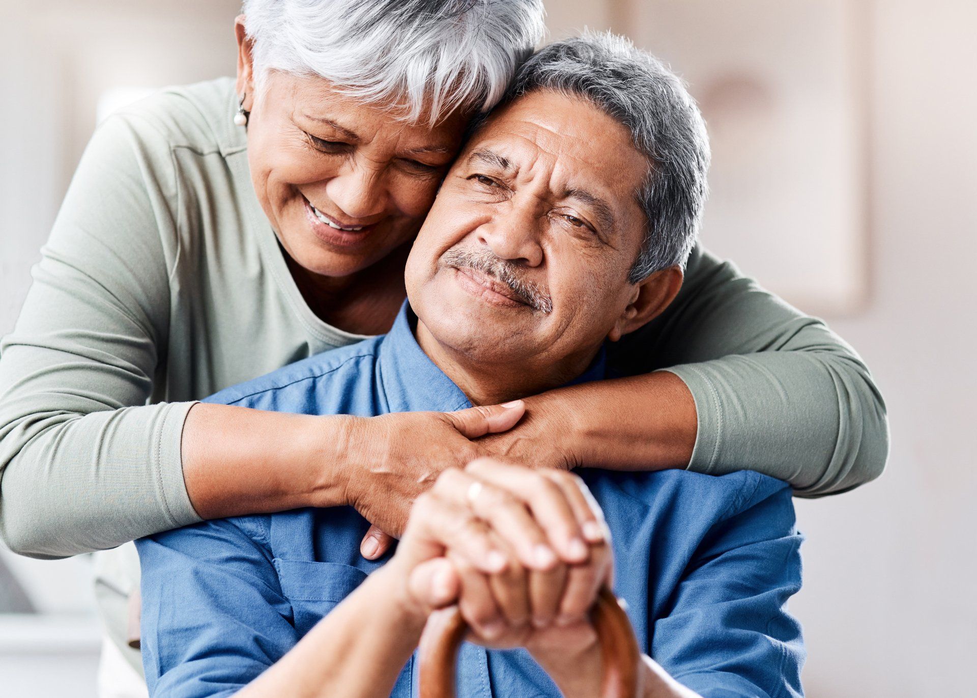 Older couple embracing; woman smiling, hugging man who holds a cane, both indoors.