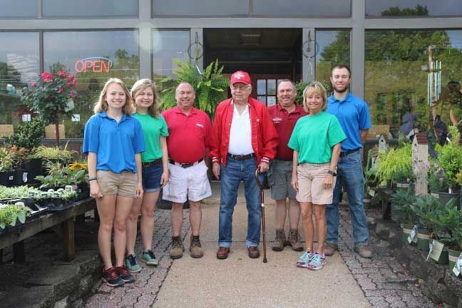 Six people stand smiling outside a garden center, with plants and an open sign behind them.