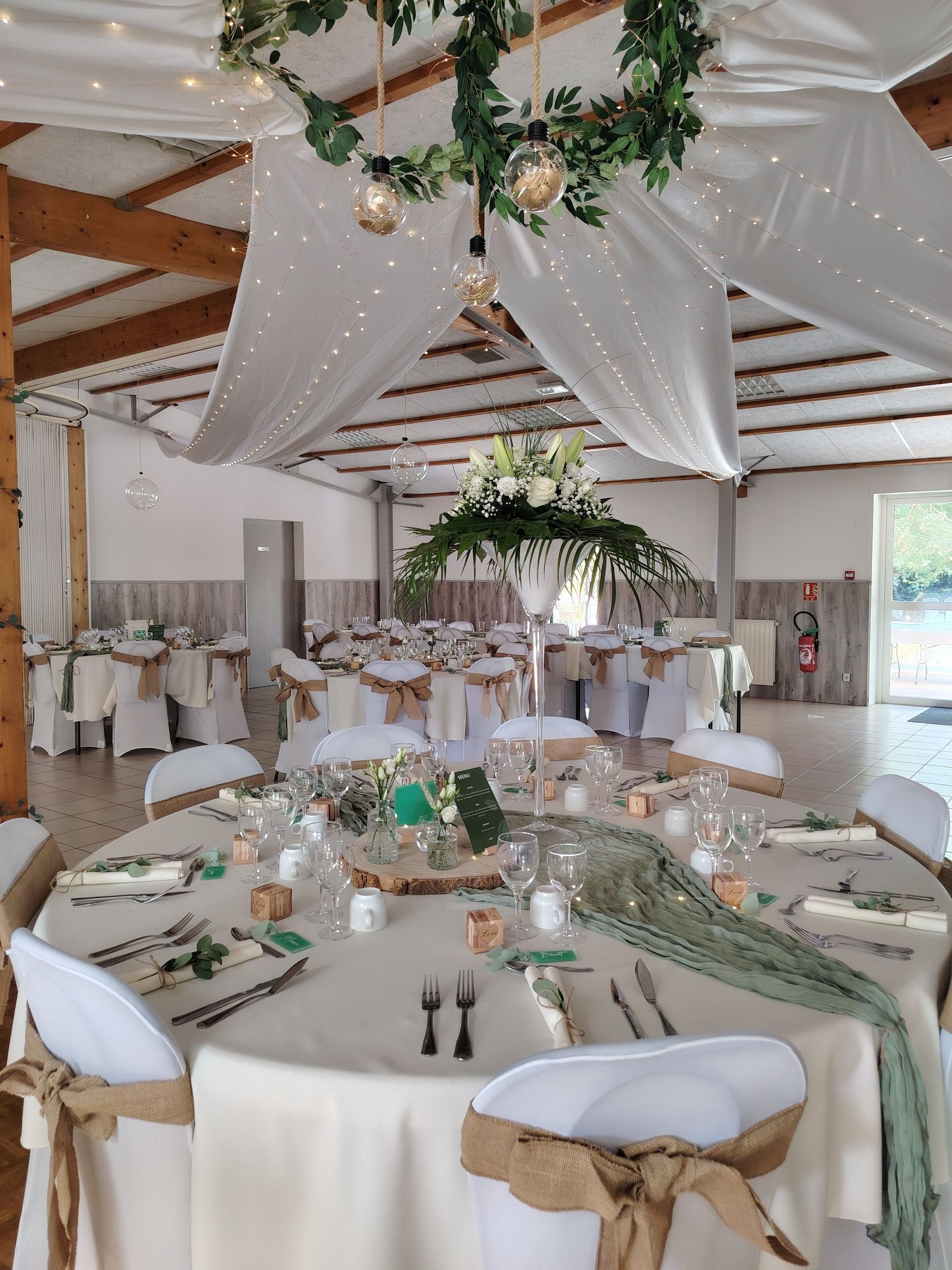 Lieu de réception de mariage élégant avec tables rondes, draperies blanches, verdure et lustres.