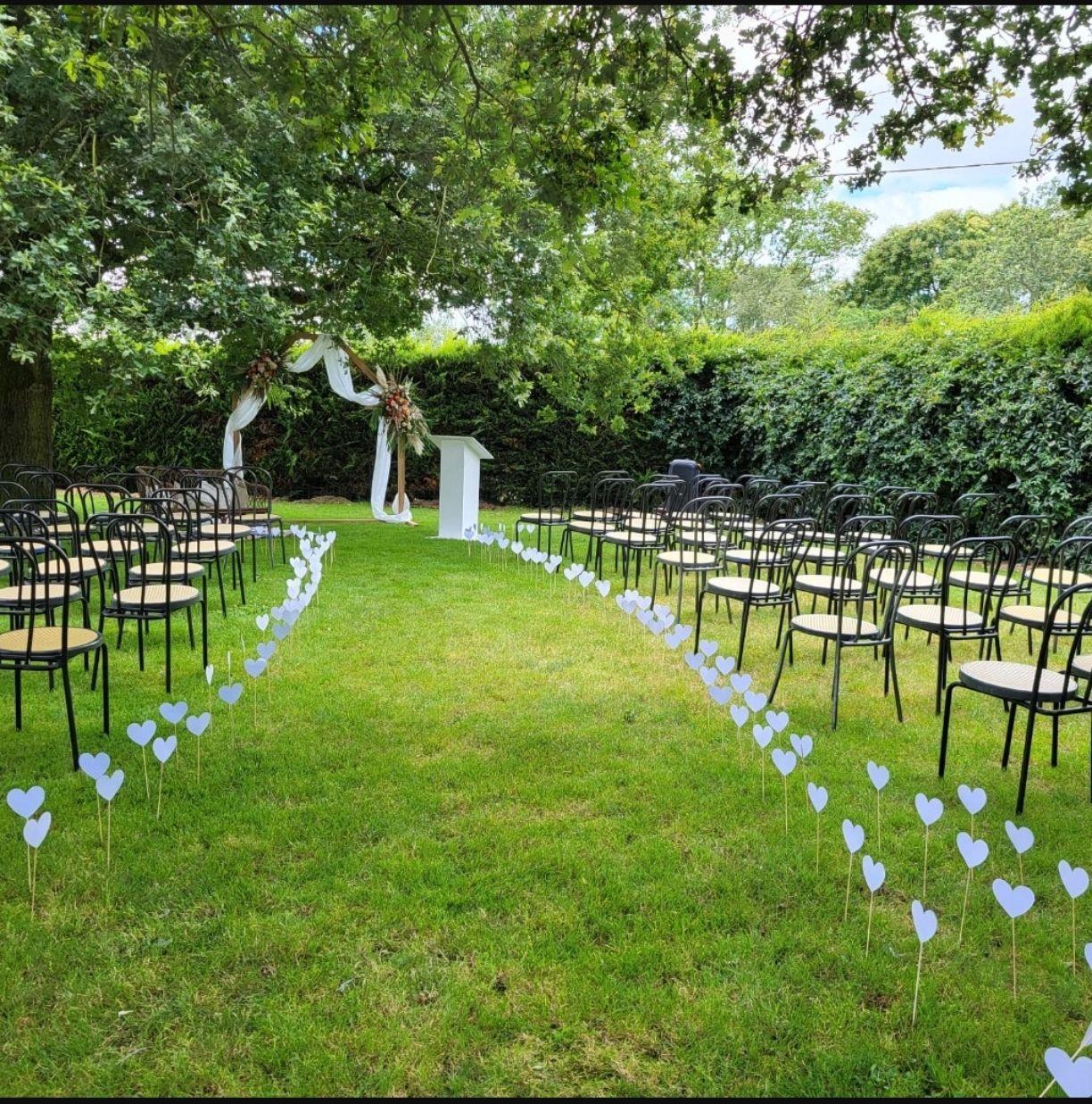 Allée nuptiale dans un jardin avec chaises noires, arche blanche et décorations en forme de cœur sous des arbres feuillus