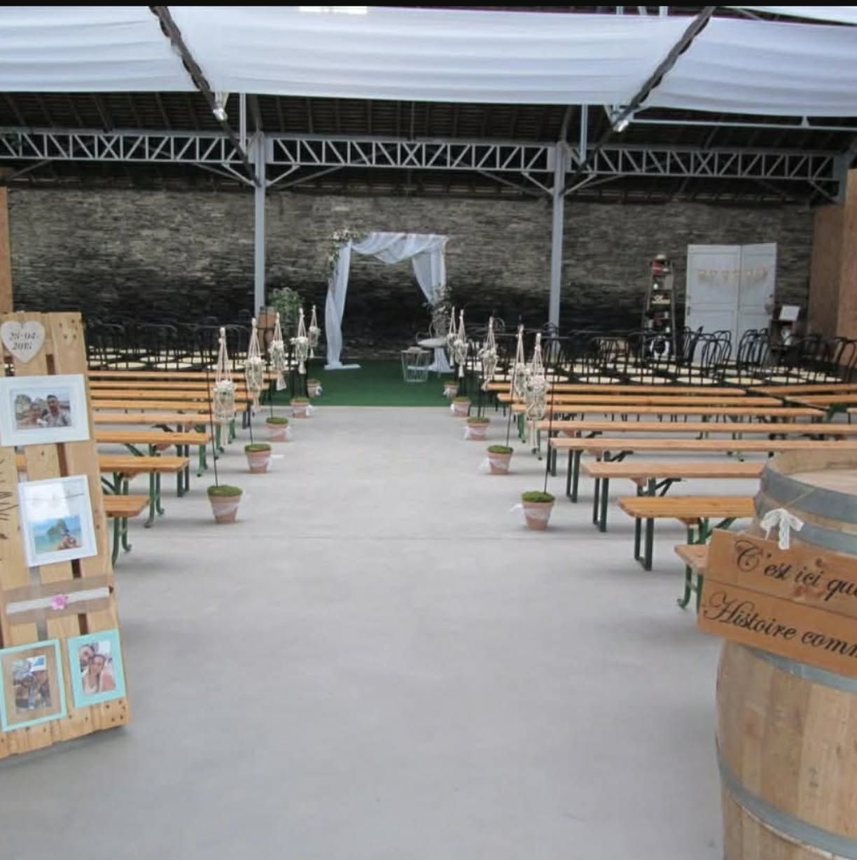 Longue allée de réception extérieure bordée de bancs en bois, menant à une arche drapée de blanc sous un pavillon couvert.