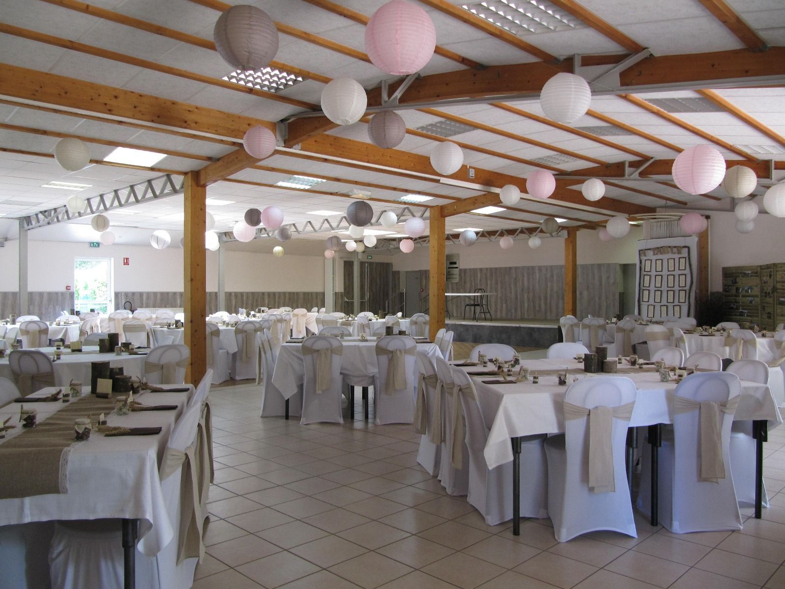Salle de banquet vide avec nappes blanches, chaises et lanternes en papier pastel suspendues sous des poutres en bois.