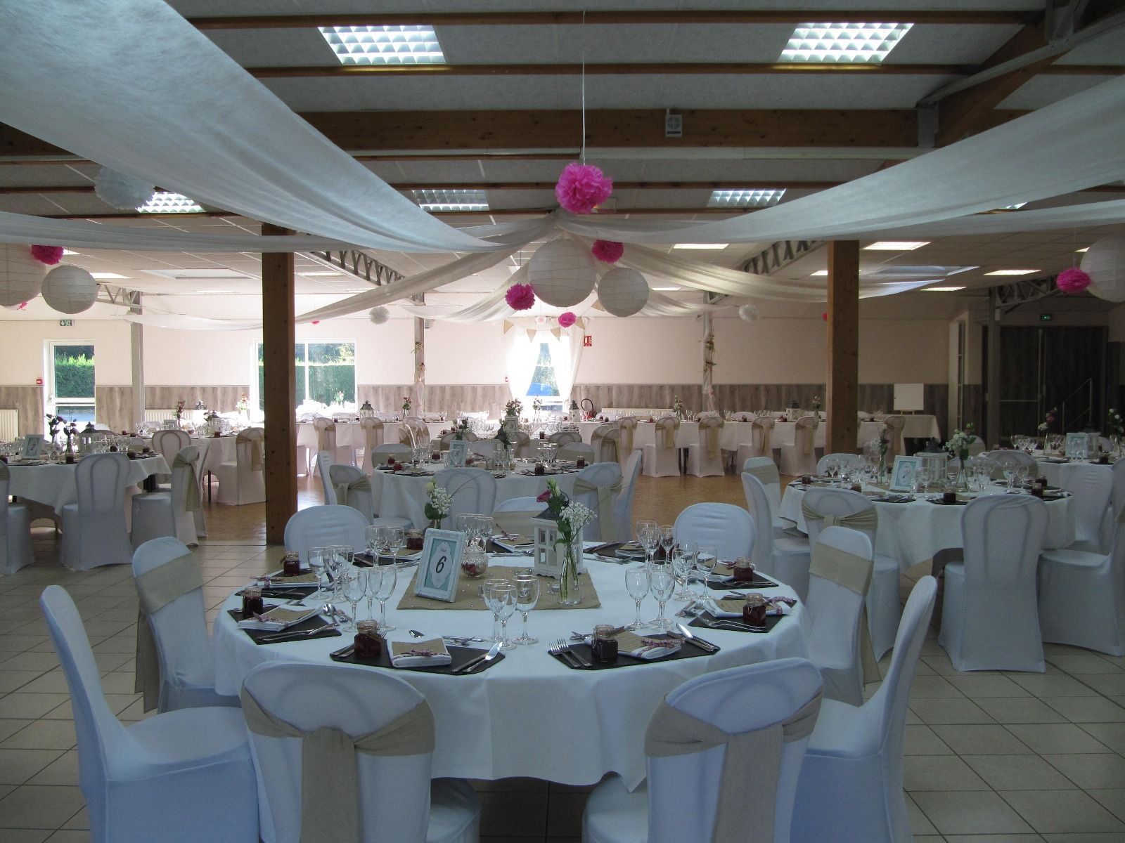 Salle de banquet aménagée avec des tables rondes, des housses de chaises blanches et des décorations suspendues roses.