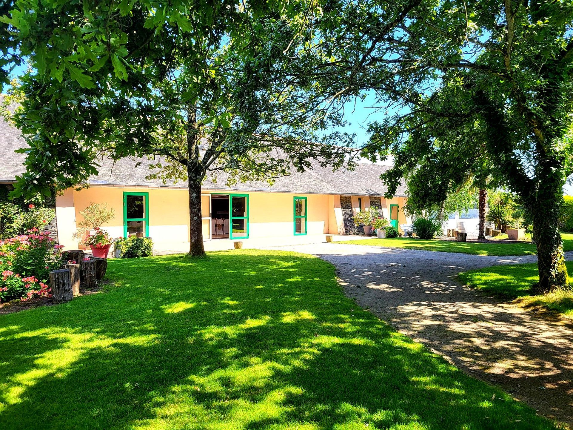 Maison de plain-pied de couleur beige avec des fenêtres à encadrement vert et une grande cour avant ombragée.