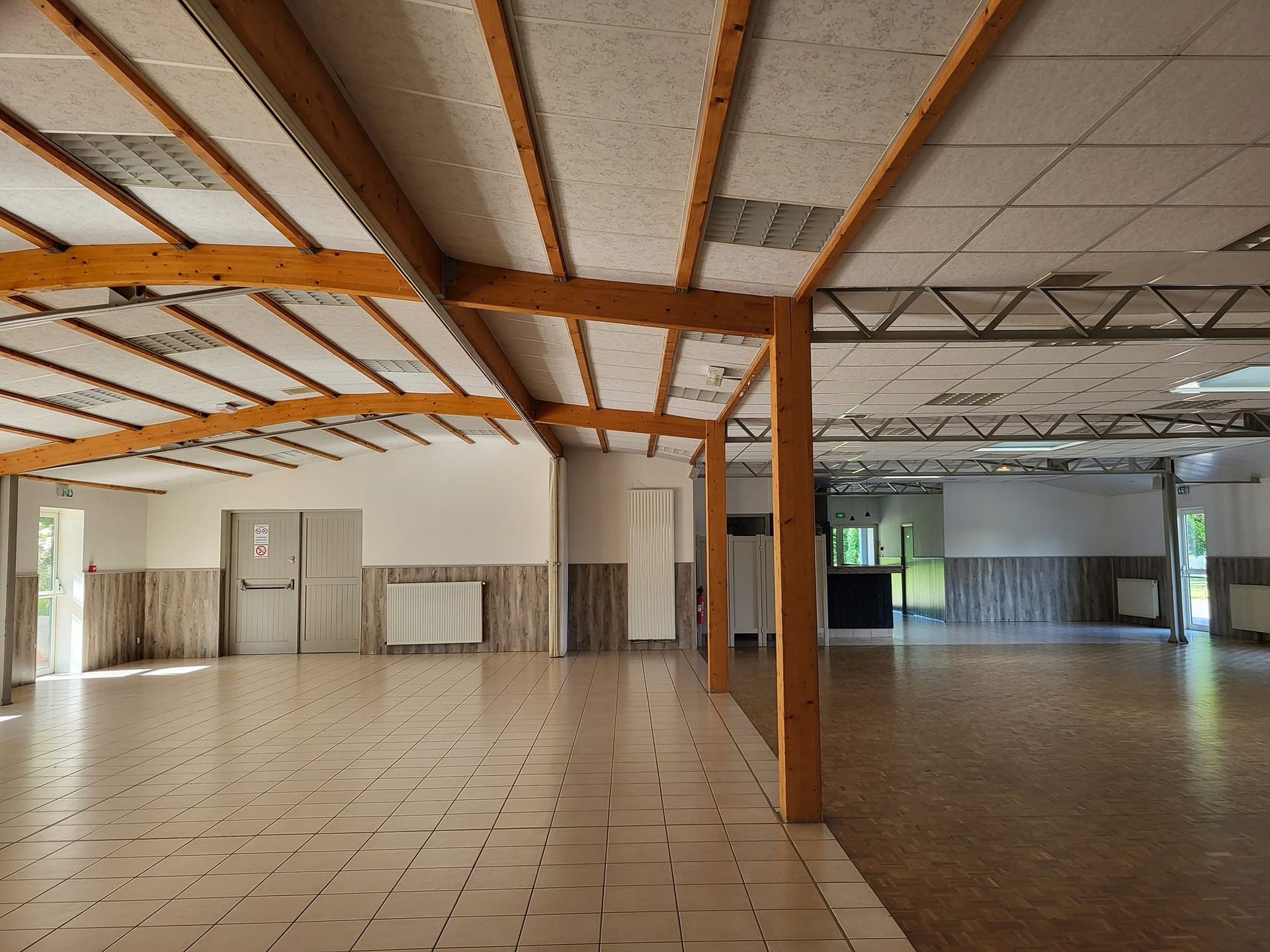 Hall intérieur vide avec sol carrelé, poutres apparentes et cloisons sous un plafond mansardé