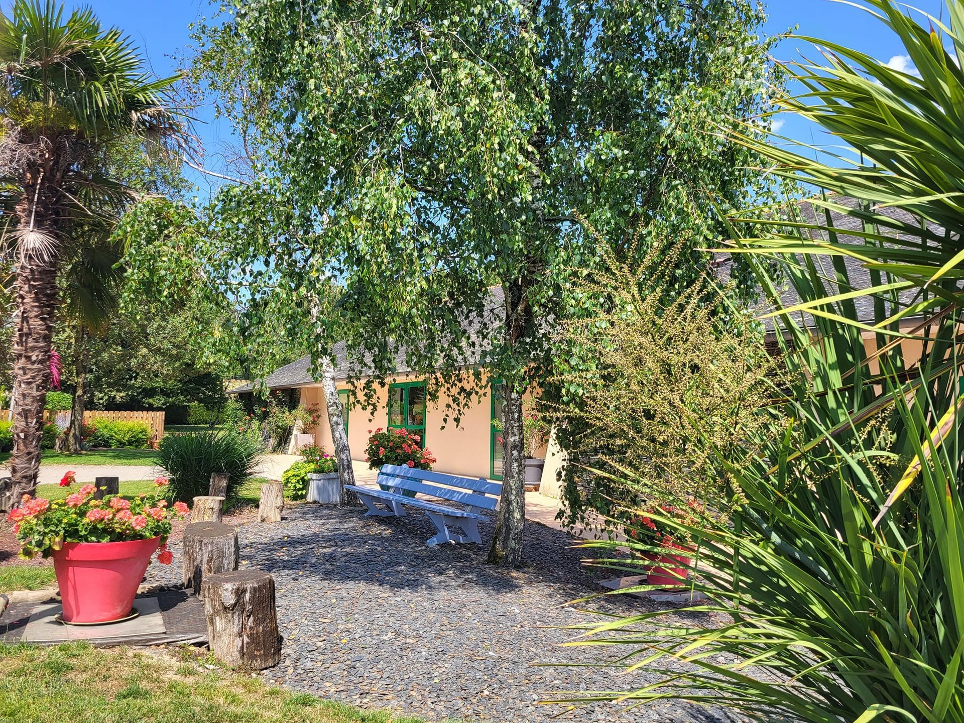 Jardin ensoleillé avec arbres, allée de gravier, pots de fleurs rouges et un petit bâtiment en arrière-plan