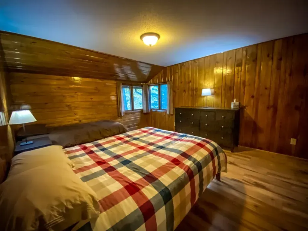 A bedroom with a bed , dresser , lamp and wooden walls.
