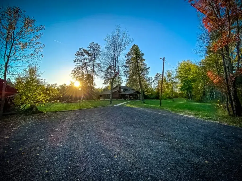 A dirt road leading to a house in the woods with the sun shining through the trees.
