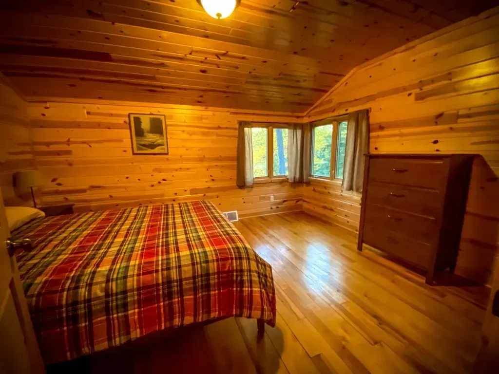 A bedroom in a log cabin with a bed , dresser , and windows.