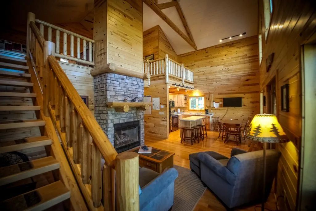 A living room in a log cabin with a fireplace and stairs leading up to the second floor.