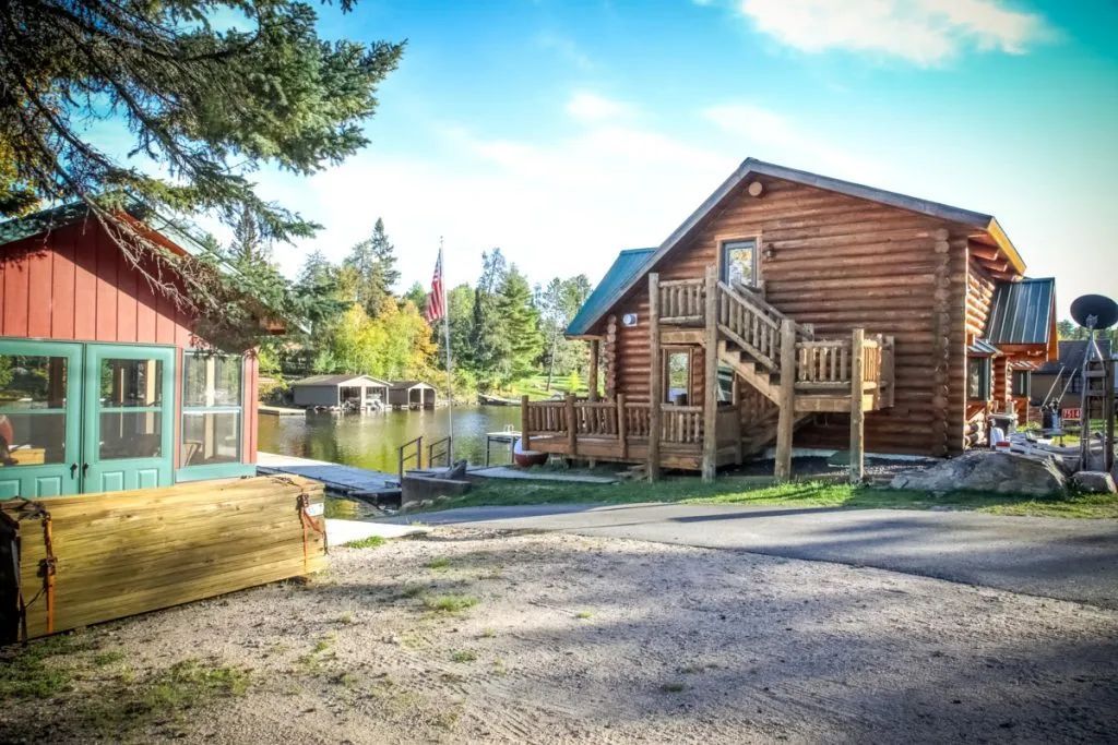 A log cabin with stairs leading up to the second floor is sitting next to a lake.