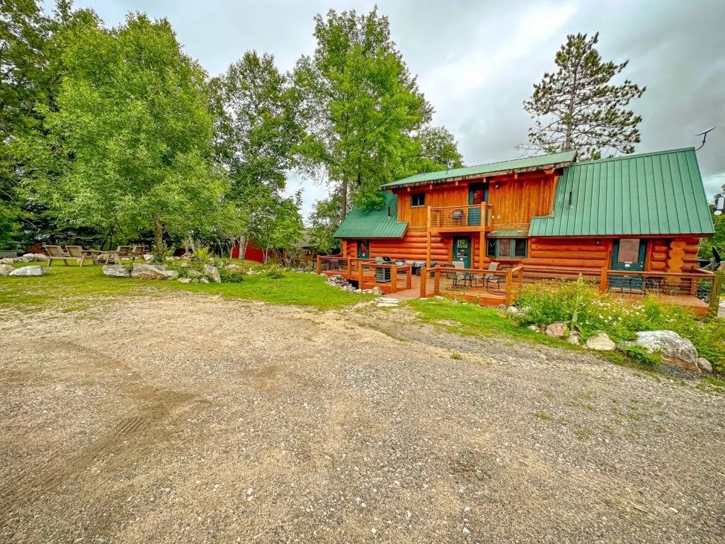 A log cabin with a green roof is surrounded by trees and a gravel driveway.