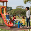 Children play on a slide and seesaw at a park while an adult watches nearby on a sunny day.