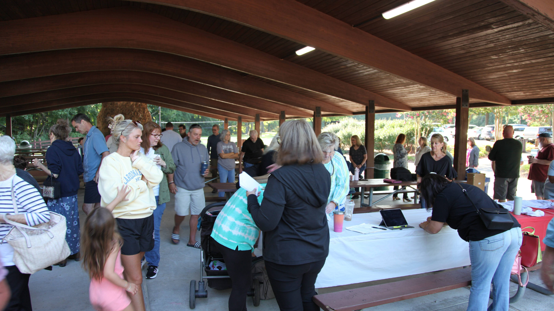 People gather at a covered outdoor event, some at a table, others conversing, in a park-like setting.