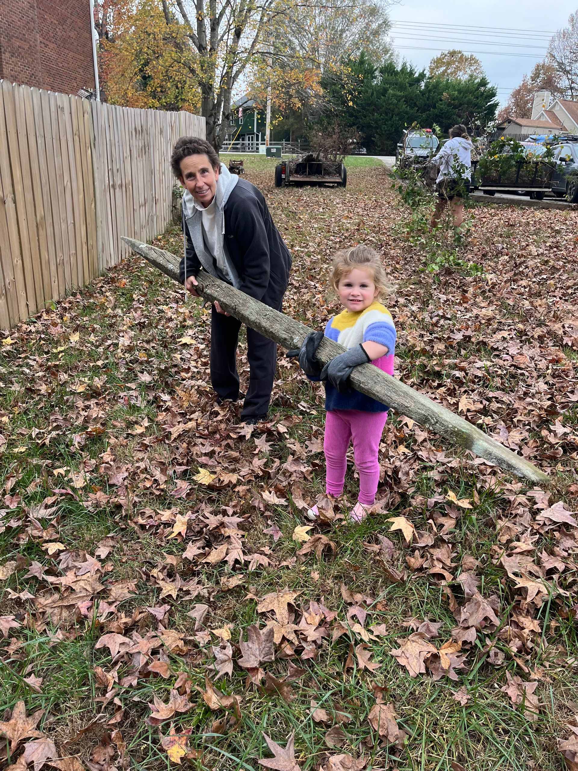 Woman and child holding a large branch in a yard covered in fallen leaves near a wooden fence.