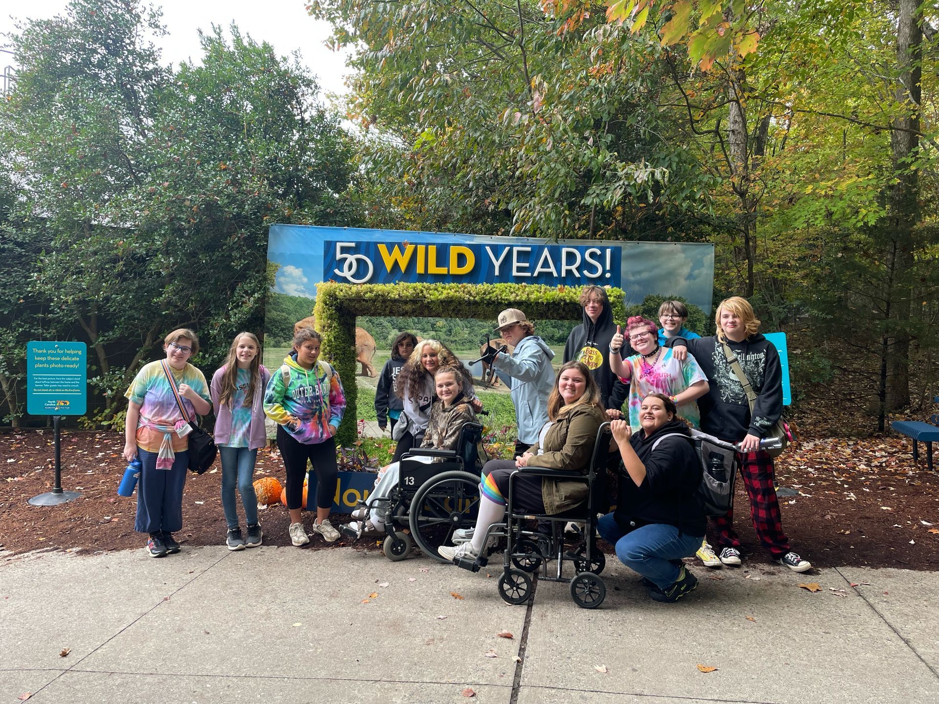 Group of people at an outdoor zoo exhibit, some in wheelchairs, posing near a