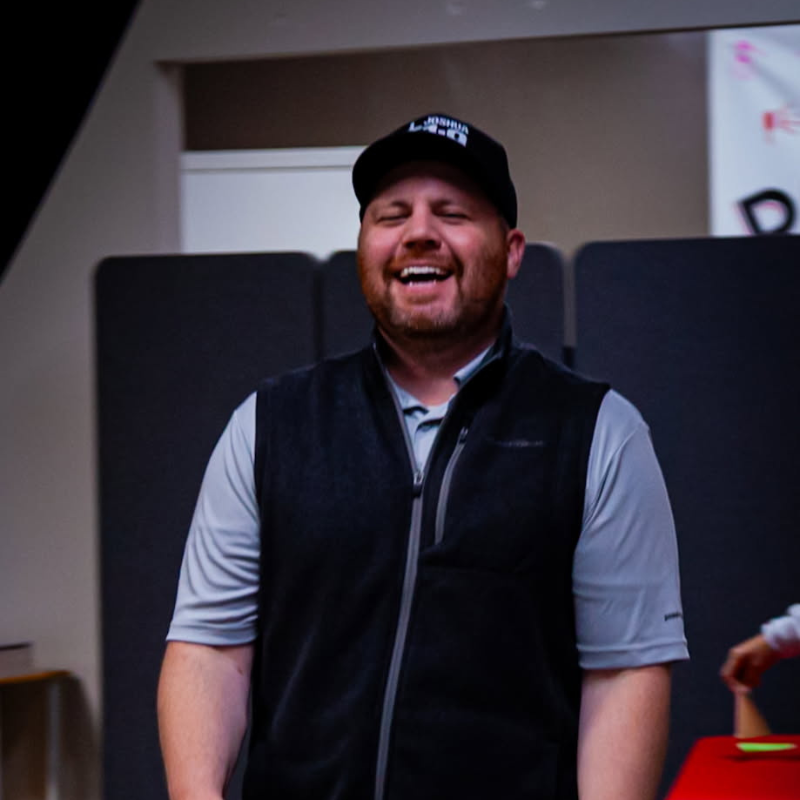 Man in a black vest and baseball cap laughs, in a room with neutral walls and dividers.