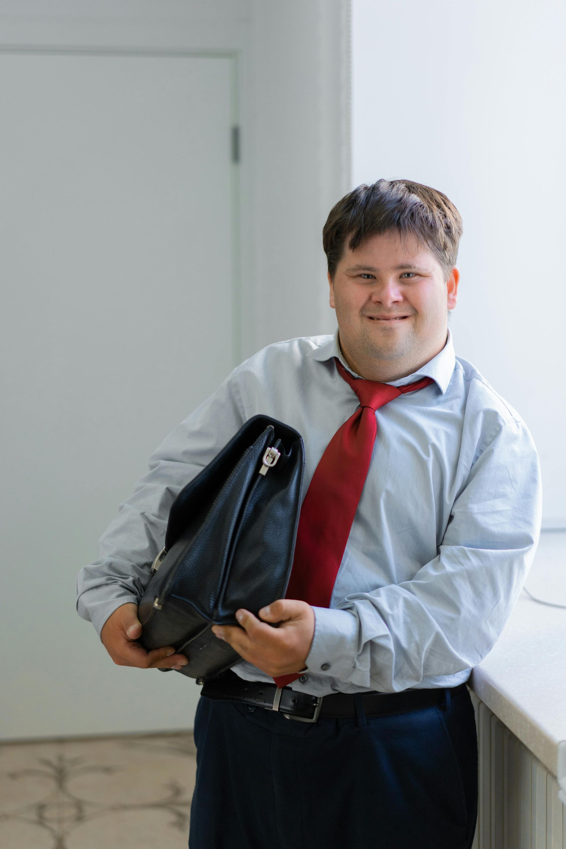 Man in business attire holding a briefcase, smiling near a window.