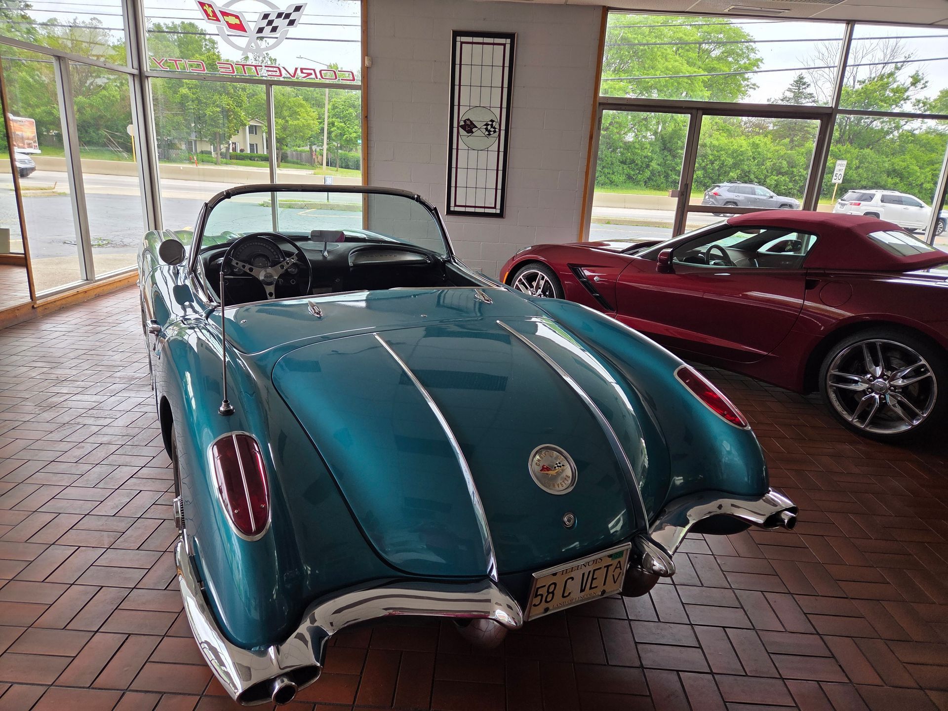 Two vintage convertibles, teal and red, displayed under a covered patio near a road