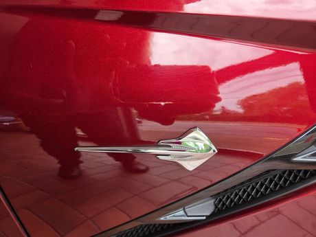 Close-up of a red car’s chrome side emblem near the front fender and vent. Close-up of a red car’s chrome side emblem near the front fender and vent.