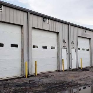 Row of white industrial garage doors on a gray warehouse, with yellow bollards and wet pavement