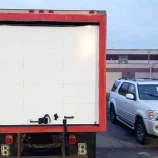 Rear of a white box truck with a red frame, parked beside a silver SUV in a lot.