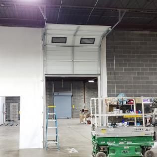 Warehouse interior with a green scissor lift, ladder, shelving, and an open loading bay.