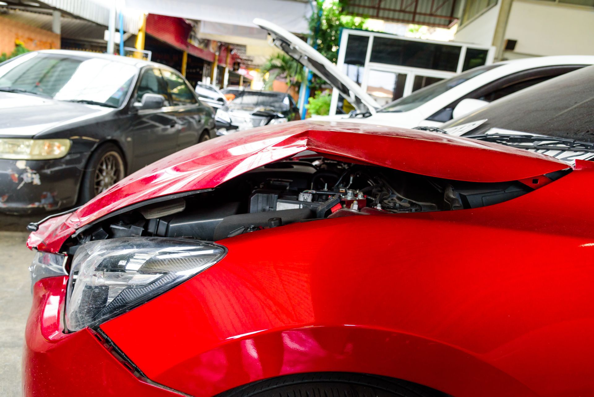 Red car with a damaged hood and front end, parked in a repair shop or garage