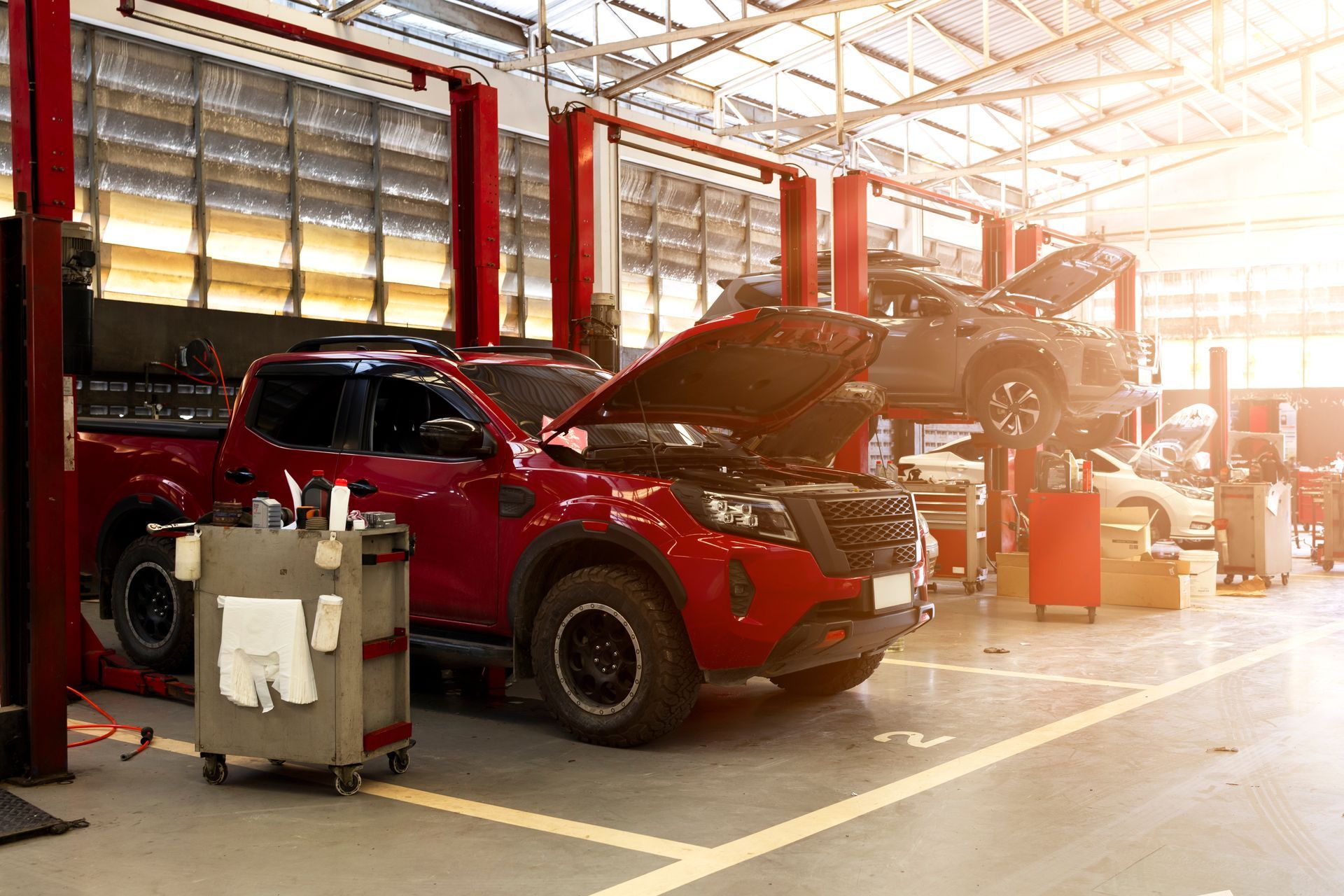 Red SUV in a brightly lit auto repair shop with its hood open and another vehicle on a lift.