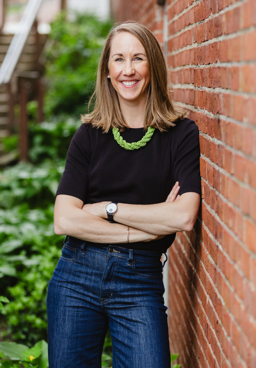 Smiling person in black shirt and jeans leaning against a brick wall outdoors