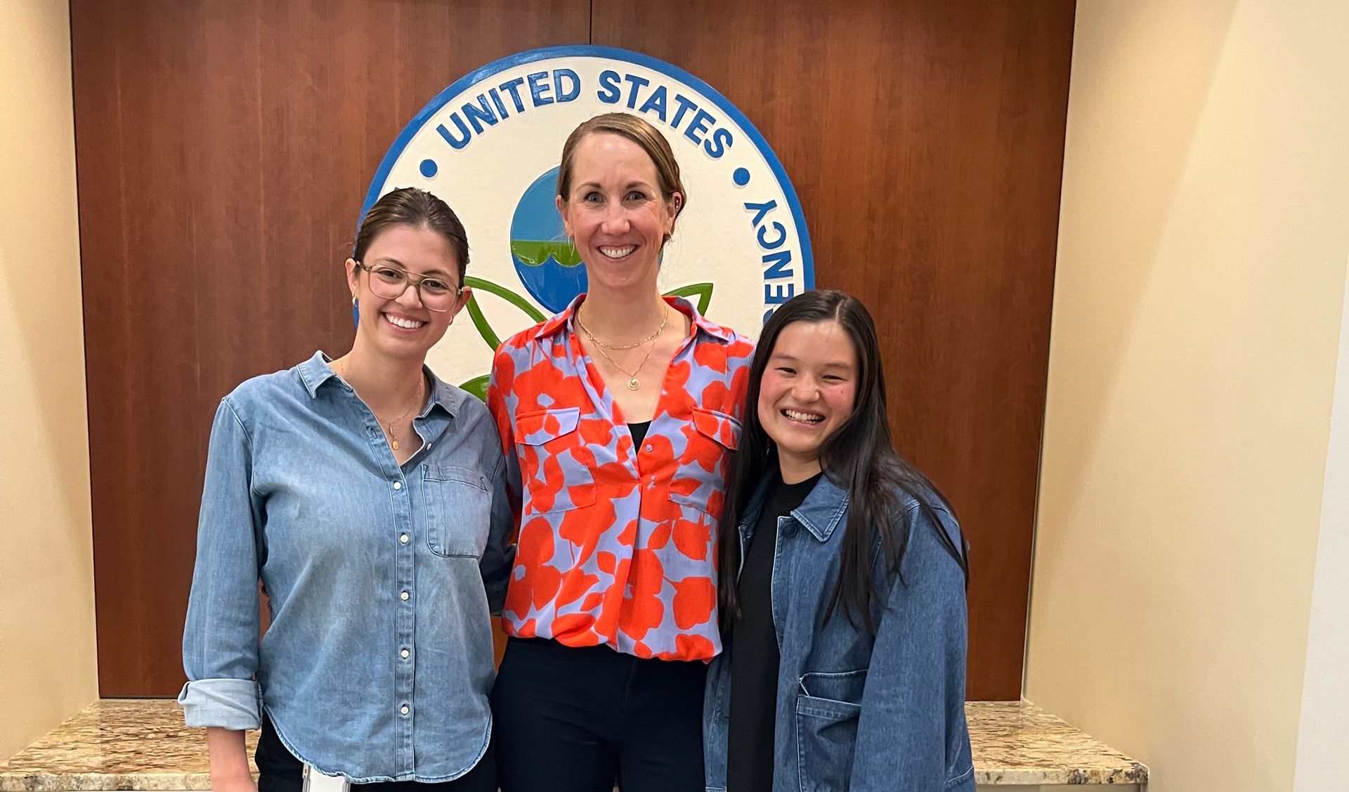 Three people standing in front of a U.S. Department of Justice seal, smiling indoors.