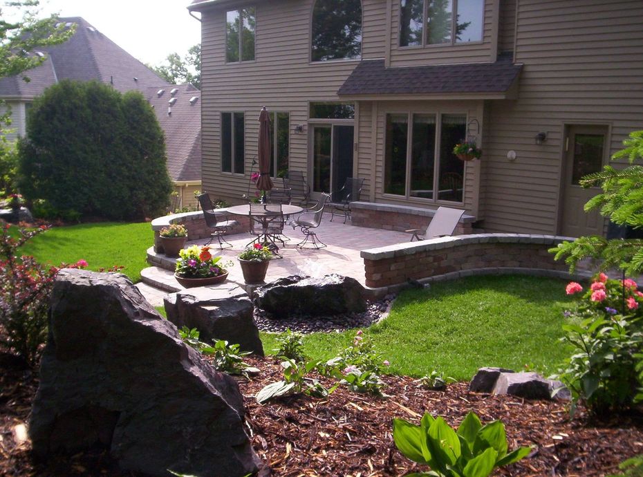 Backyard patio with wooden table and chairs, stone retaining wall, and green lawn behind a house