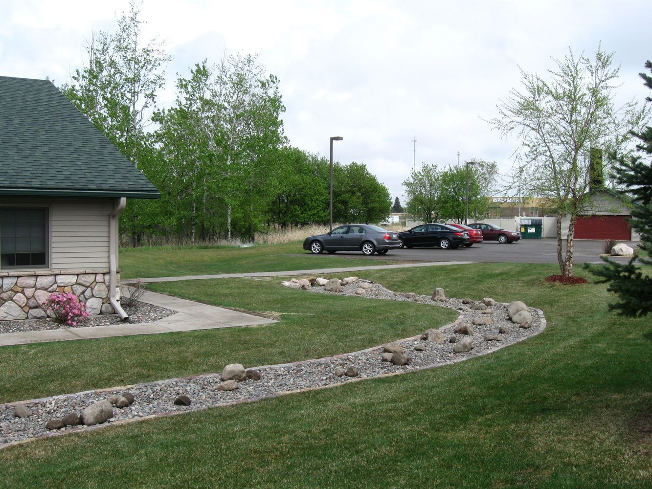 Curved rock garden edging a grassy yard beside a house, with parked cars and trees in the background.