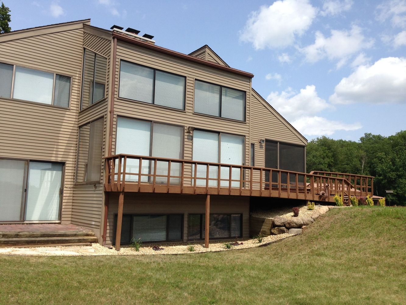 Two-story beige house with large windows, wooden deck, and grassy yard under a blue sky