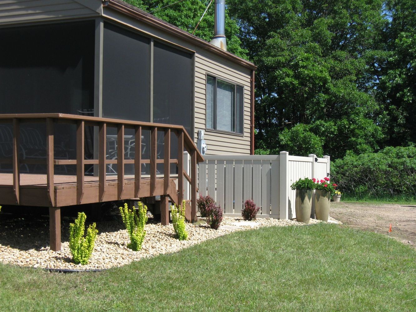Mobile home porch with wood railing, small garden, and gravel yard in a sunny wooded setting