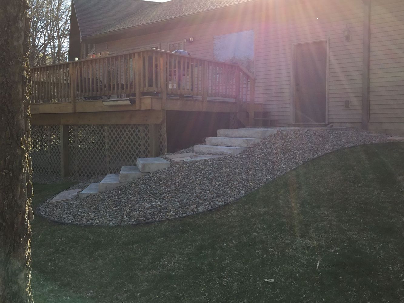 Wooden deck with steps over gravel slope beside a house, sun glare in the background.