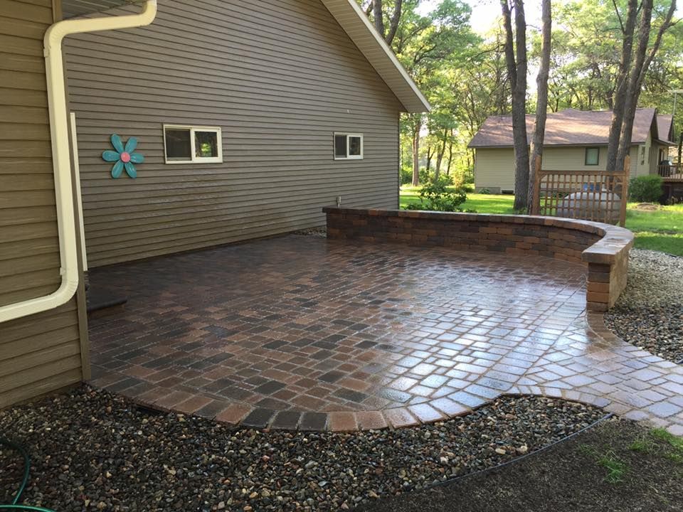 Brick-paver patio beside a house with a low retaining wall and wooded backyard.