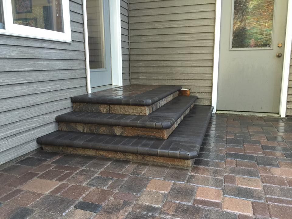 Front porch with three black steps, gray siding, and a tiled patio beside a door and window.