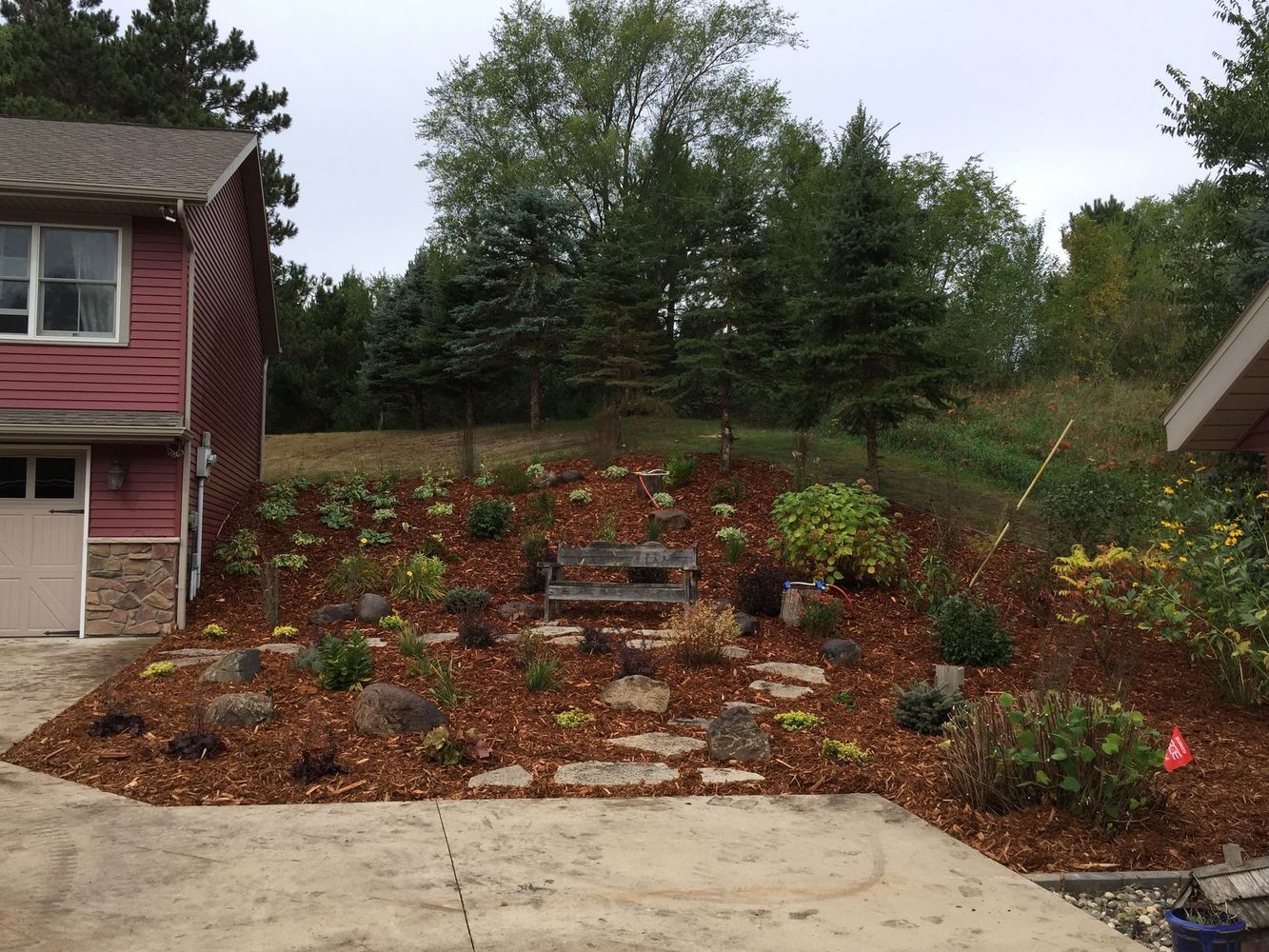 Front yard garden bed with stone steps, mulch, and shrubs beside a house, viewed from a driveway